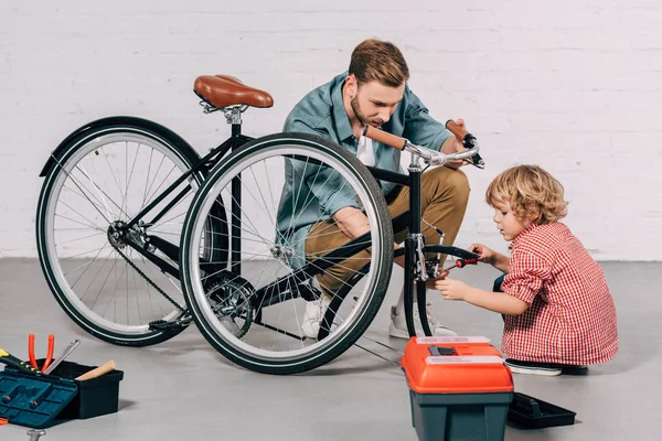 Hombre ayudar a niño pequeño fijación de bicicleta cerca de herramientas caja en taller - foto de stock