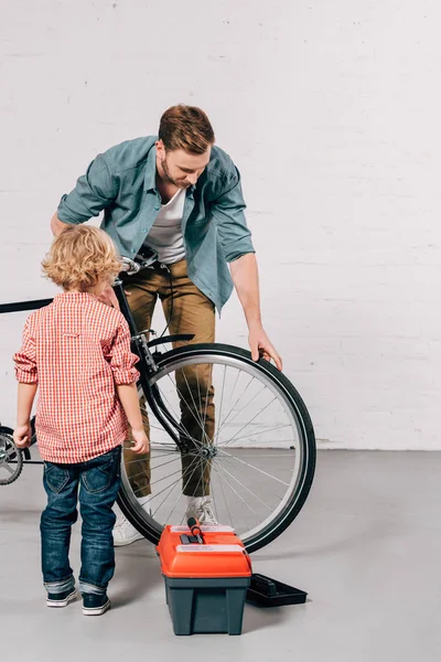 Padre mostrando pequeño hijo cómo fijar bicicleta cerca de herramientas caja en taller - foto de stock