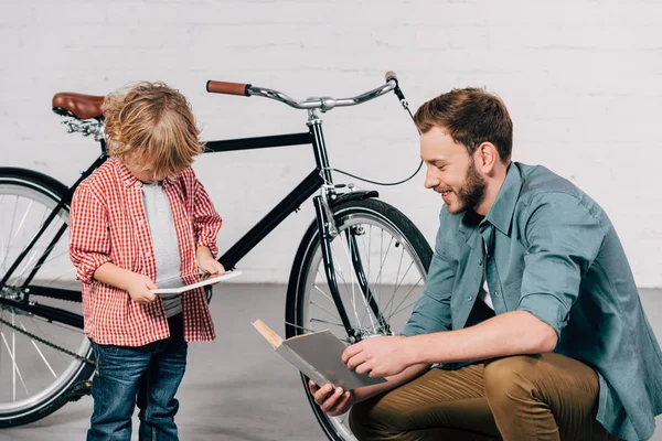 Junge nutzt digitales Tablet, während Vater neben Fahrrad Buch liest — Stockfoto