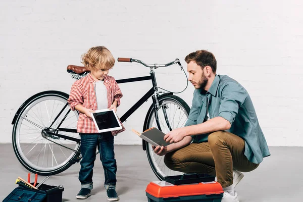 Kleines Kind mit digitalem Tablet und Vater liest Buch in Fahrradwerkstatt — Stockfoto