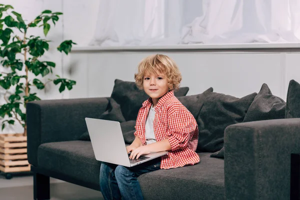 Kleines Kind schaut in die Kamera und benutzt Laptop auf dem heimischen Sofa — Stockfoto