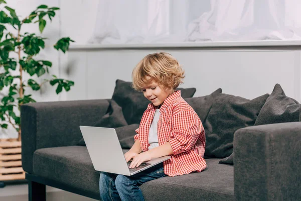 Menino encaracolado feliz usando laptop no sofá em casa — Fotografia de Stock
