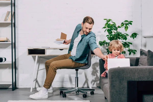 Kleiner Junge mit Laptop auf Sofa, während sein Vater zu Hause mit der Hand auf Bildschirm zeigt — Stockfoto