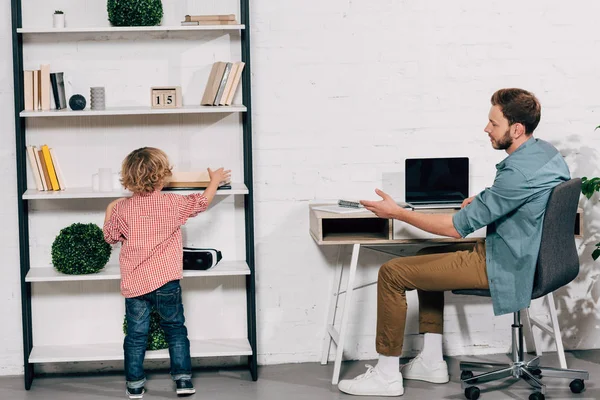 Mann zeigt mit der Hand auf Sohn, während sein Buch zu Hause im Regal steht — Stockfoto