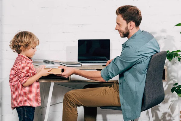 Seitenansicht eines Mannes, der seinem kleinen Sohn ein Buch am heimischen Tisch mit Laptop schenkt — Stockfoto