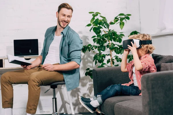 Happy sitting with book at table and looking at son using virtual reality headset on sofa at home — Stock Photo