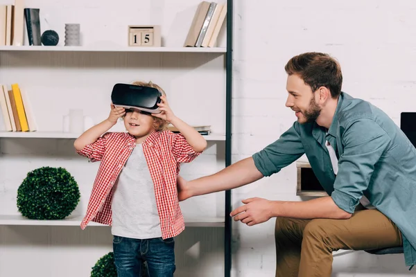 Кучерявий хлопчик одягає гарнітуру віртуальної реальності, а його батько сидить поруч вдома — стокове фото