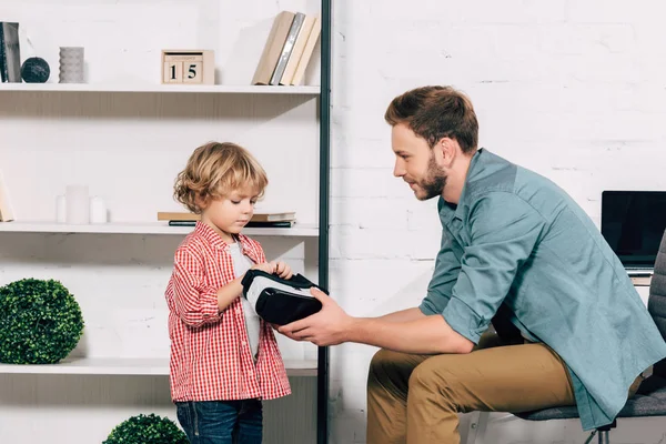 Side view of man giving virtual reality headset to adorable little son at home — Stock Photo