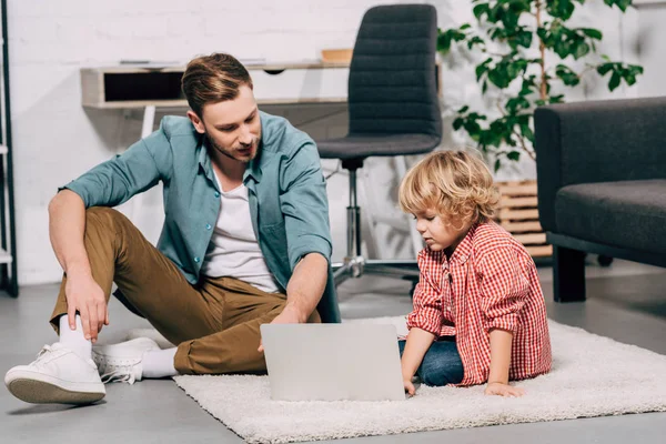 Pai e filho sentados no chão com laptop em casa — Fotografia de Stock