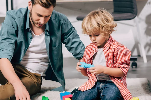 Vista de cerca del niño y su padre jugando con bloques de plástico de colores en el suelo en casa - foto de stock