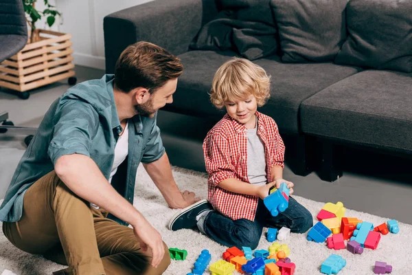 Vista de ángulo alto de niño feliz y su padre jugando con bloques de plástico de colores en el suelo en casa - foto de stock