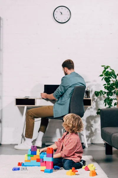 Rückansicht eines kleinen Jungen, der mit Plastikklötzen auf dem Boden spielt, während sein Vater zu Hause am Laptop arbeitet — Stockfoto