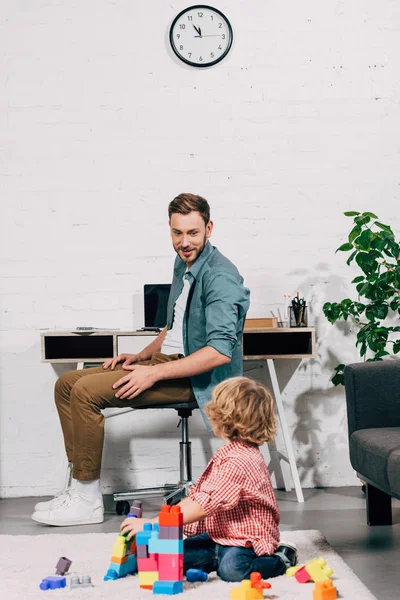 Boy playing with plastic blocks and talking to father sitting behind at home — Stock Photo