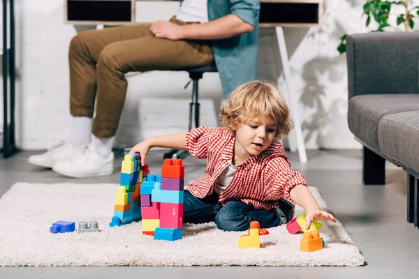 Lockiger kleiner Junge, der mit Plastikklötzen auf dem Boden spielt, während sein Vater zu Hause hinten sitzt — Stockfoto