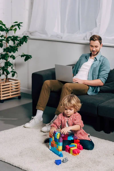 Vista de ángulo alto de niño jugando con bloques de plástico en el suelo, mientras que su padre usando el ordenador portátil en el sofá en casa - foto de stock