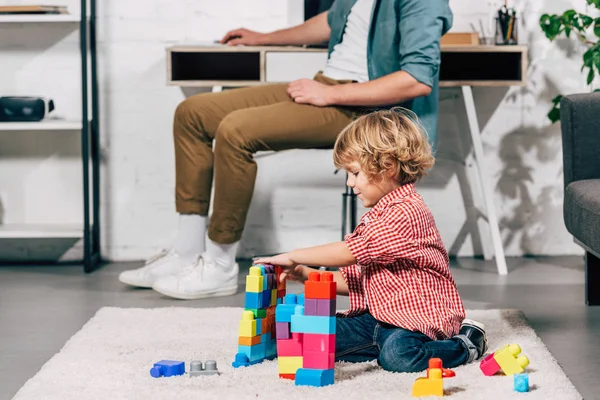 Seitenansicht eines Kindes, das mit Plastikklötzen auf dem Boden spielt, während sein Vater zu Hause hinten sitzt — Stockfoto