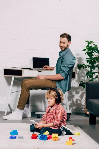 Enfoque selectivo de niño en auriculares escuchando música con teléfono inteligente y sentado en el suelo cerca de bloques de plástico, mientras que su padre sentado detrás de casa - foto de stock