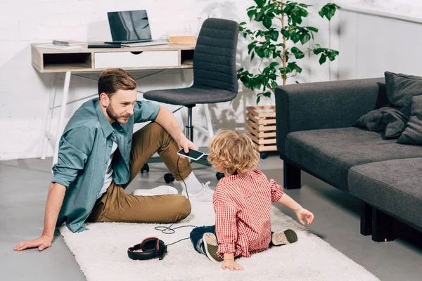 Vista trasera del niño sentado en el suelo con el padre sosteniendo el teléfono inteligente con auriculares en casa - foto de stock