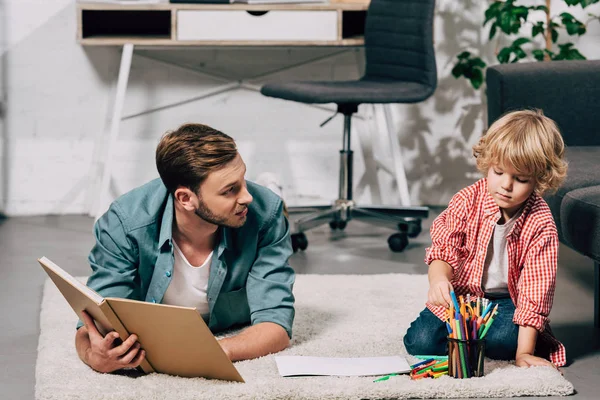 Vater liest Buch, während sein Sohn zu Hause mit bunten Filzstiften zeichnet — Stockfoto