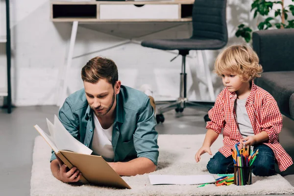 Joven hombre leyendo libro mientras su pequeño hijo dibujo con marcadores de colores en el suelo en casa - foto de stock