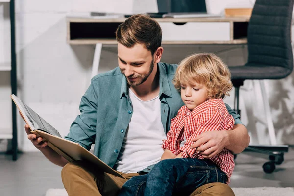 Sonriente hombre sosteniendo pequeño hijo de rodillas y leyendo libro para él - foto de stock