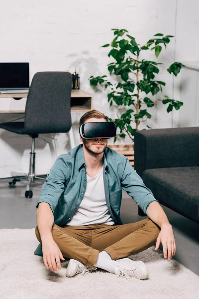 Jeune homme utilisant casque de réalité virtuelle et assis sur le sol à la maison — Photo de stock