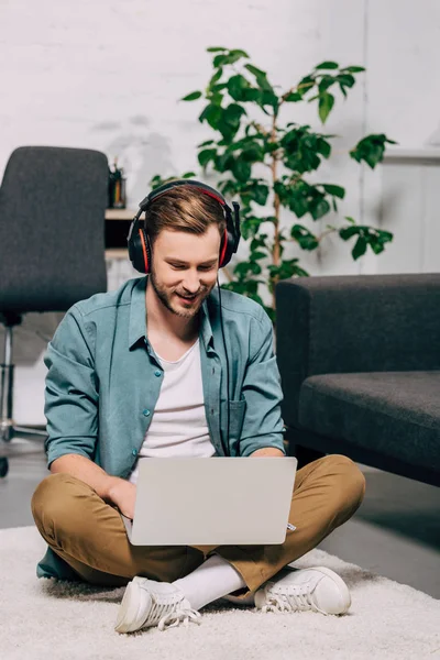 Freelancer masculino sorridente em fones de ouvido trabalhando no laptop enquanto sentado no chão em casa — Fotografia de Stock