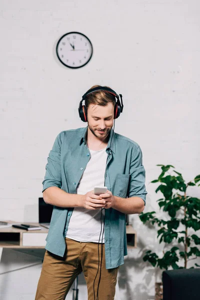 Fröhlicher junger Mann mit Kopfhörern, der Musik hört und das Smartphone benutzt — Stockfoto