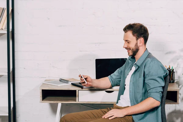 Vista lateral del freelancer masculino feliz usando el smartphone en la mesa con el ordenador portátil - foto de stock