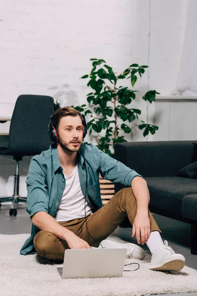 Jovem freelancer masculino em fones de ouvido trabalhando no laptop enquanto sentado no chão em casa — Fotografia de Stock