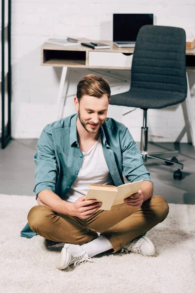 Glücklicher junger Mann sitzt auf dem Boden und liest zu Hause Buch — Stockfoto