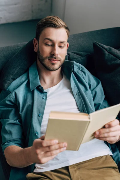 Vue grand angle du jeune homme allongé sur le canapé et le livre de lecture à la maison — Photo de stock