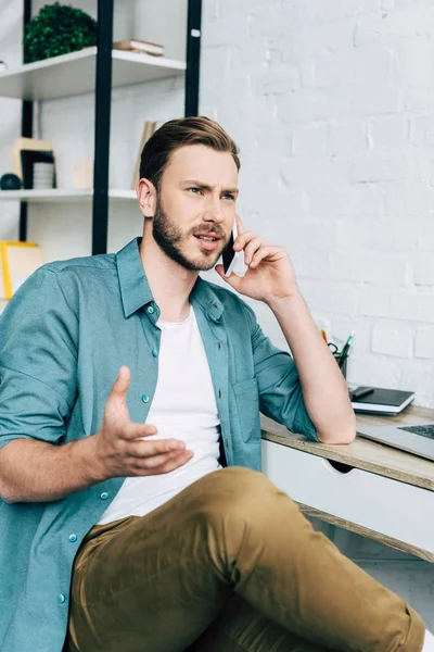 Irritierter junger männlicher Freiberufler spricht am Tisch mit Smartphone — Stockfoto