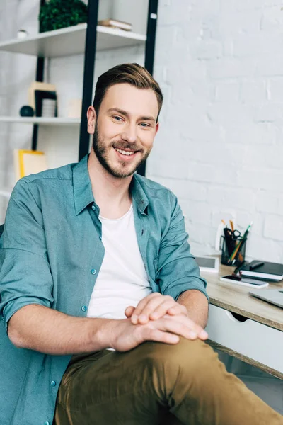 Sonriente joven mirando a la cámara mientras está sentado cerca de la mesa - foto de stock