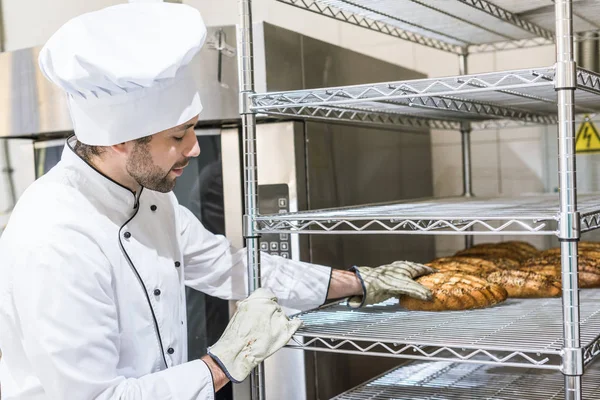 Panadero masculino adulto tocando pan recién horneado en estante de cocina - foto de stock