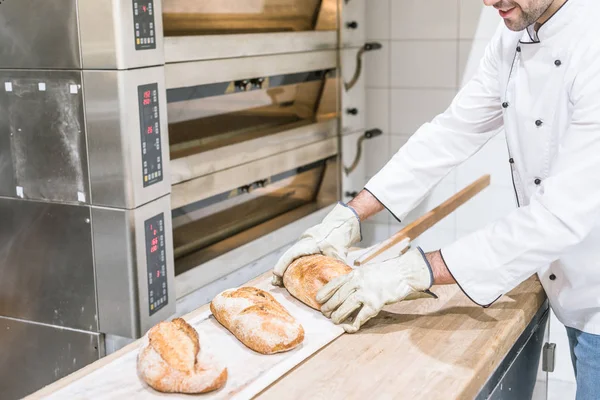 Panettiere in piedi vicino al forno con pane caldo cotto sul bancone di legno — Foto stock