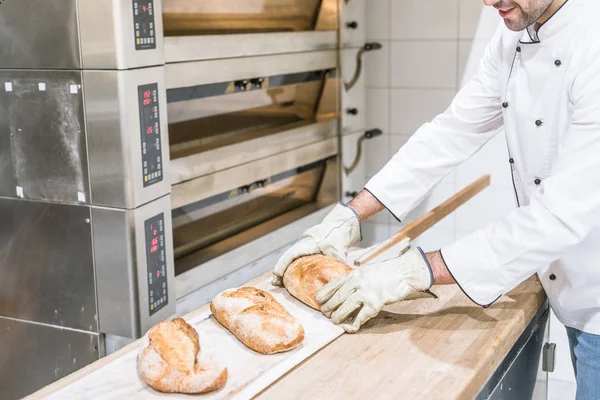 Panadero de pie cerca del horno con pan caliente horneado en el mostrador de madera - foto de stock