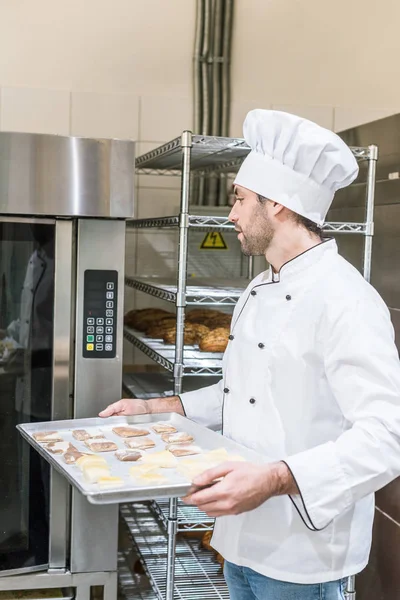 Adult baker in white chefs uniform putting baking tray with uncooked dough in oven — Stock Photo