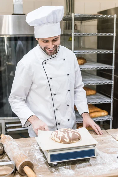 Smiling baker weighing raw dough on kitchen scales — Stock Photo