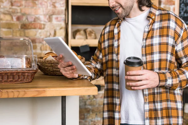 Обрізаний вид людини за допомогою цифрового планшета і тримає одноразову чашку з кавою в пекарні — стокове фото