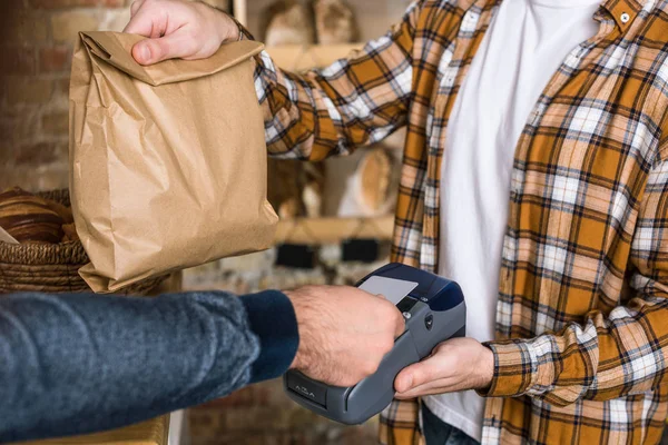 Close up do vendedor segurando saco de papel enquanto o cliente paga pela compra com cartão de crédito na padaria — Fotografia de Stock