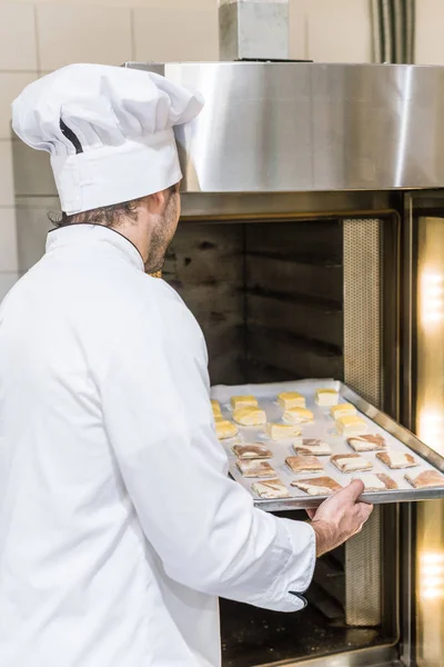 Male baker in white chefs uniform putting baking tray with uncooked dough in oven — Stock Photo