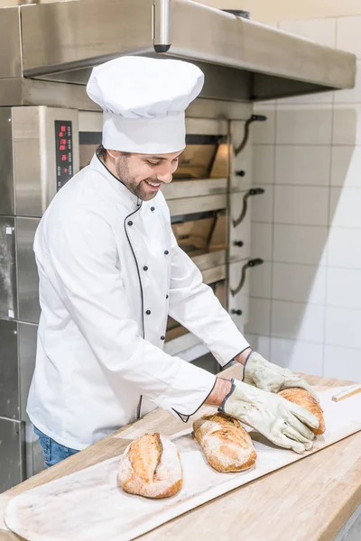 Smiling baker standing near oven with fresh baked bread on wooden counter — Stock Photo