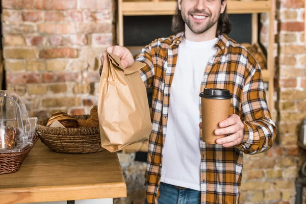 Sonriente hombre sosteniendo bolsa de papel con pastelería y café para ir a la panadería - foto de stock