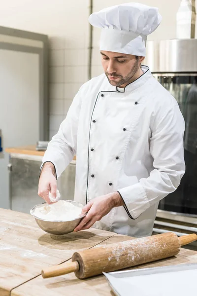 Panadero masculino adulto dispersando harina en tablero de madera en la cocina - foto de stock