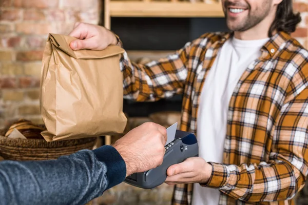 Abgeschnittene Ansicht eines lächelnden Verkäufers, der Papiertüte hält, während der Kunde mit Kreditkarte bezahlt — Stockfoto