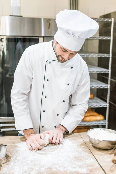Cook in white chefs uniform kneading dough on wooden table — Stock Photo