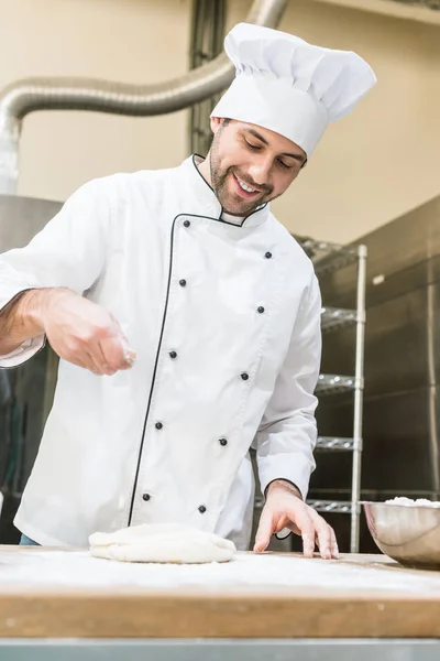 Panadero en uniforme de chefs blancos sonriendo y cocinando masa en cocina profesional - foto de stock
