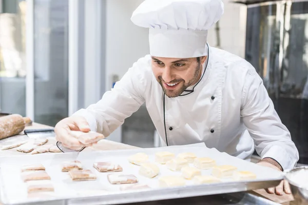 Panadero en uniforme de chefs sonriendo y poniendo masa sin cocer en bandeja - foto de stock