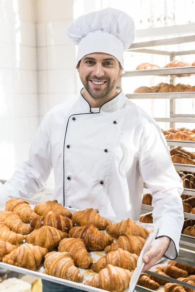 Panadero sonriente en uniforme de chefs blancos con bandeja llena de cruasanes frescos - foto de stock